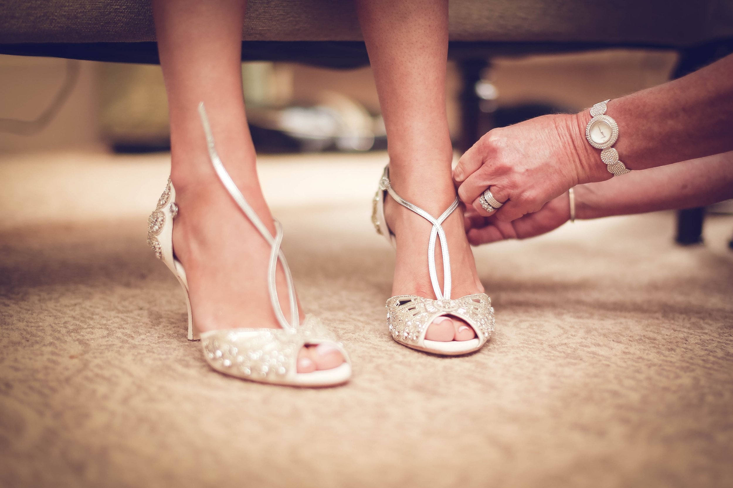Close-up of a person helping another person put on sparkly high-heeled shoes on carpeted floor.