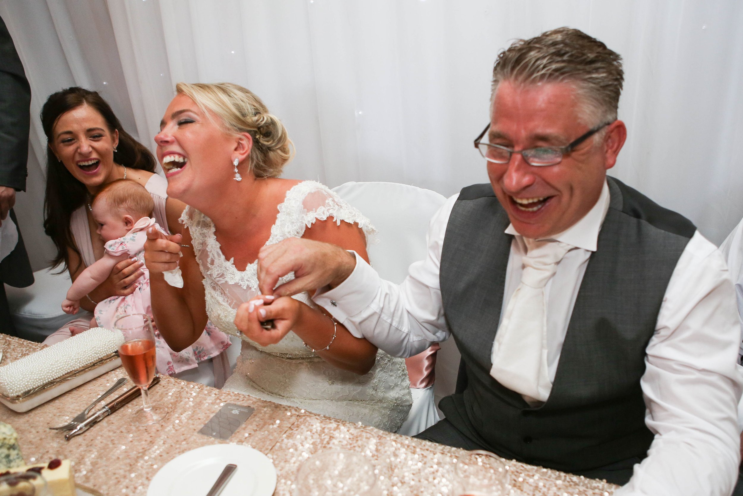 People laughing at a wedding reception with a bride, a man in a vest, and a woman holding a baby.