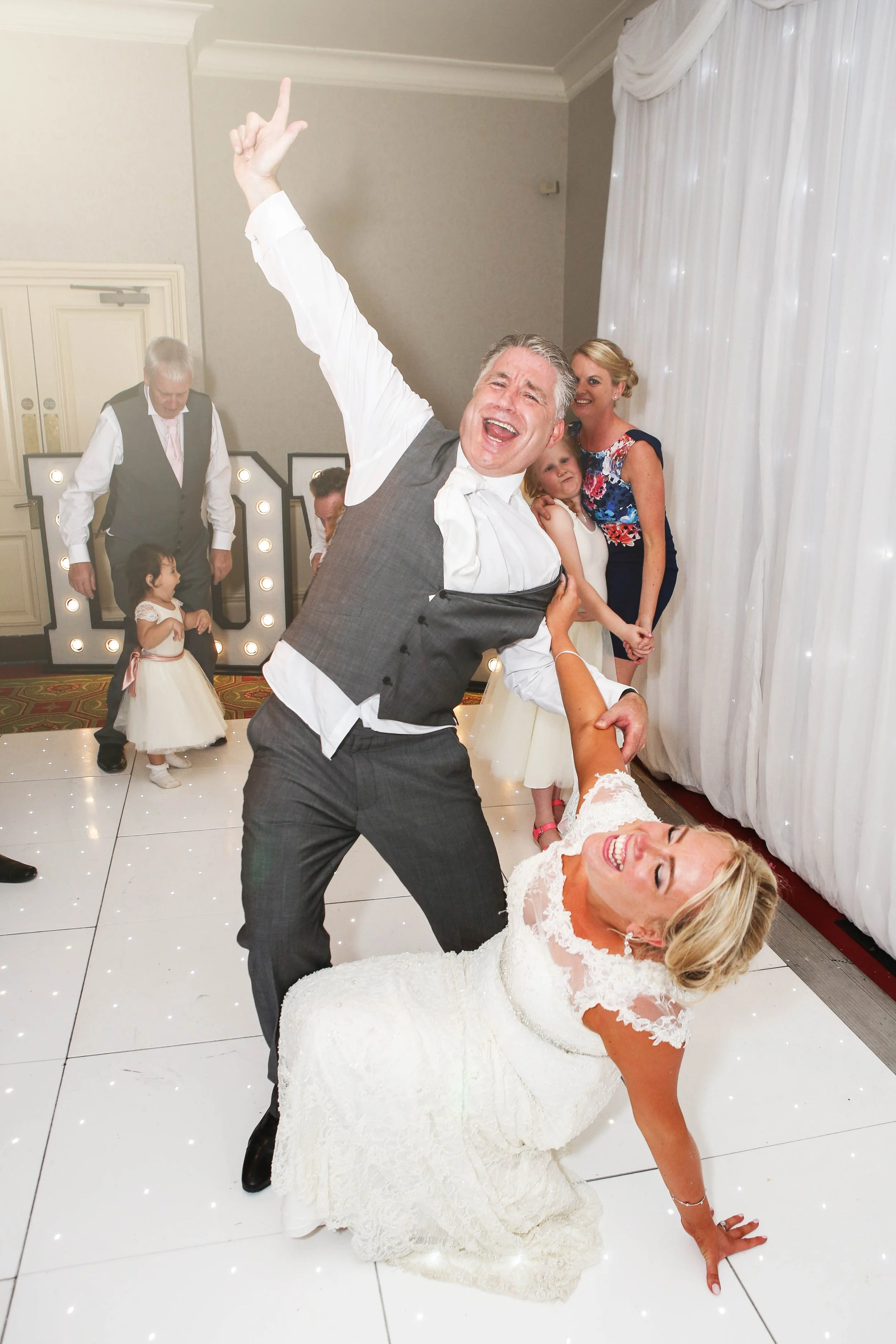 Bride and groom laughing and dancing at wedding reception with guests and children in background.