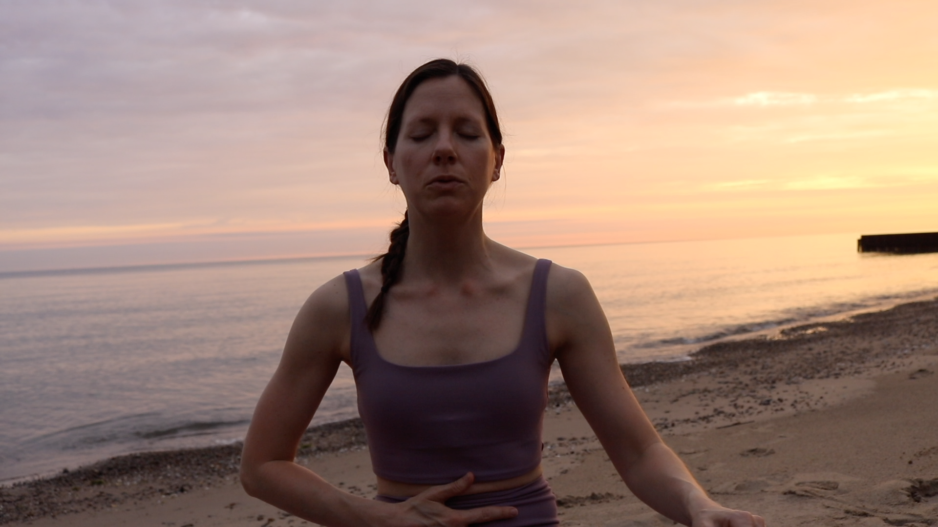 A woman with long auburn hair in a braid practicing Breathwork on a beach during sunrise, wearing a purple tank top.