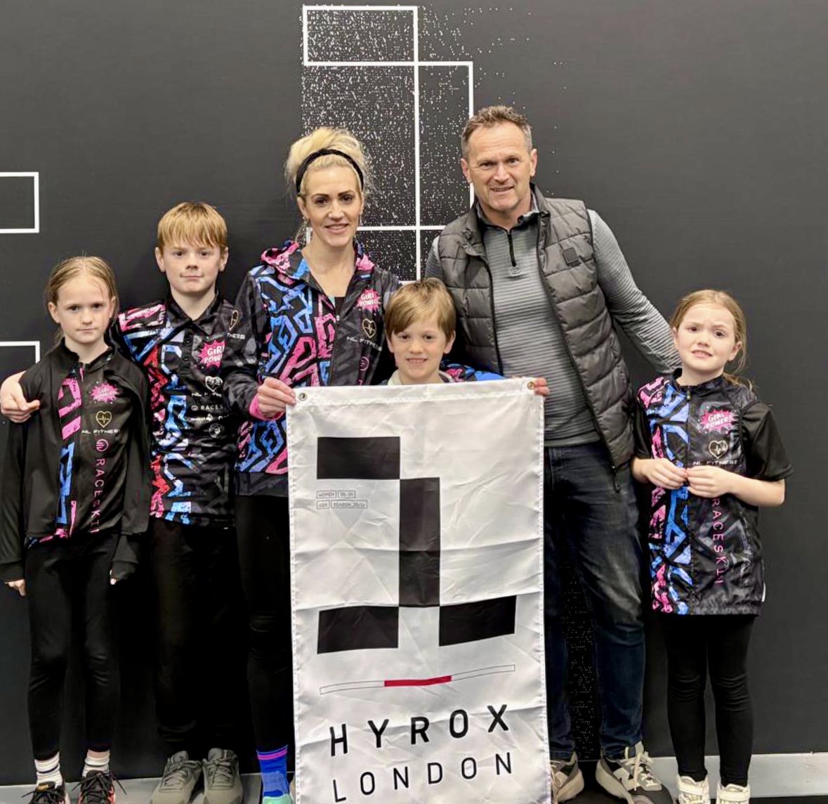 Nat Lawrence standing with her family and holding a HYROX London flag after competition.