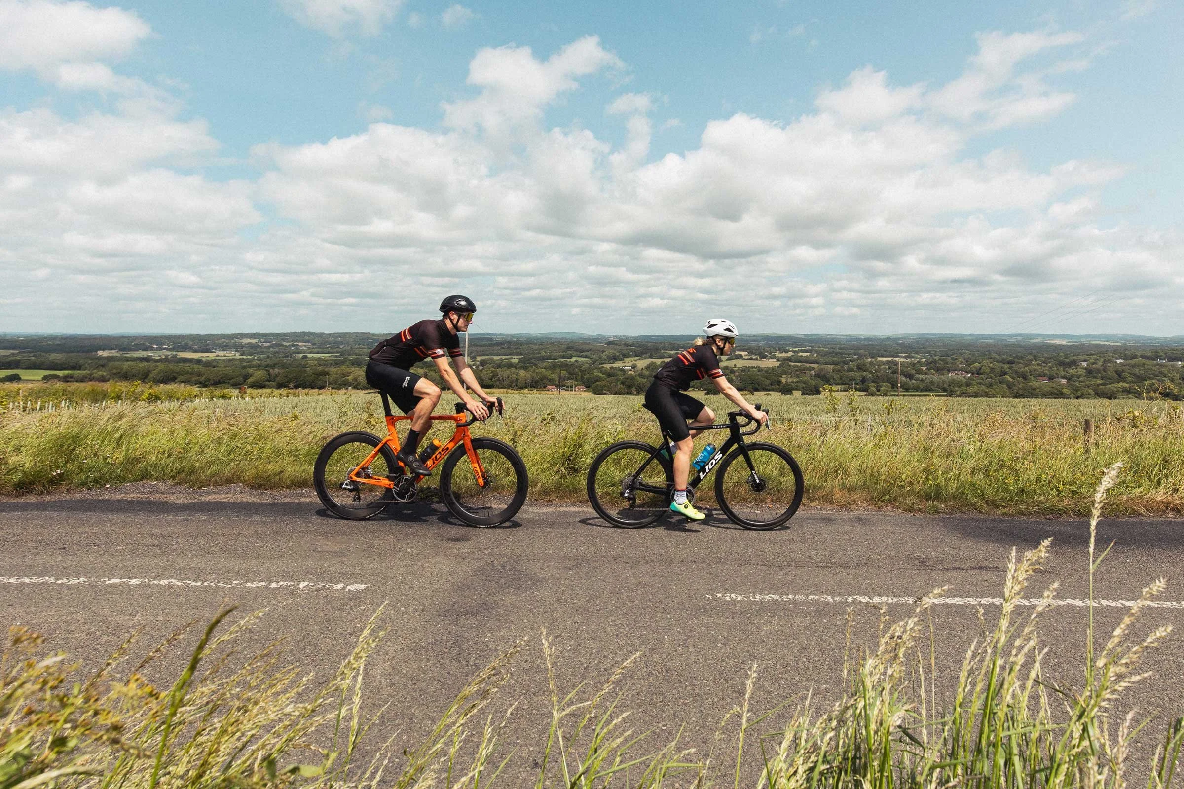 Two LIOS cyclists riding left to right on a country lane.  The first cyclist is a female riding a LIOS Bullet 5.56 endurance road bike.  The second cyclist is a male riding a LIOS Exactor aero road bike.