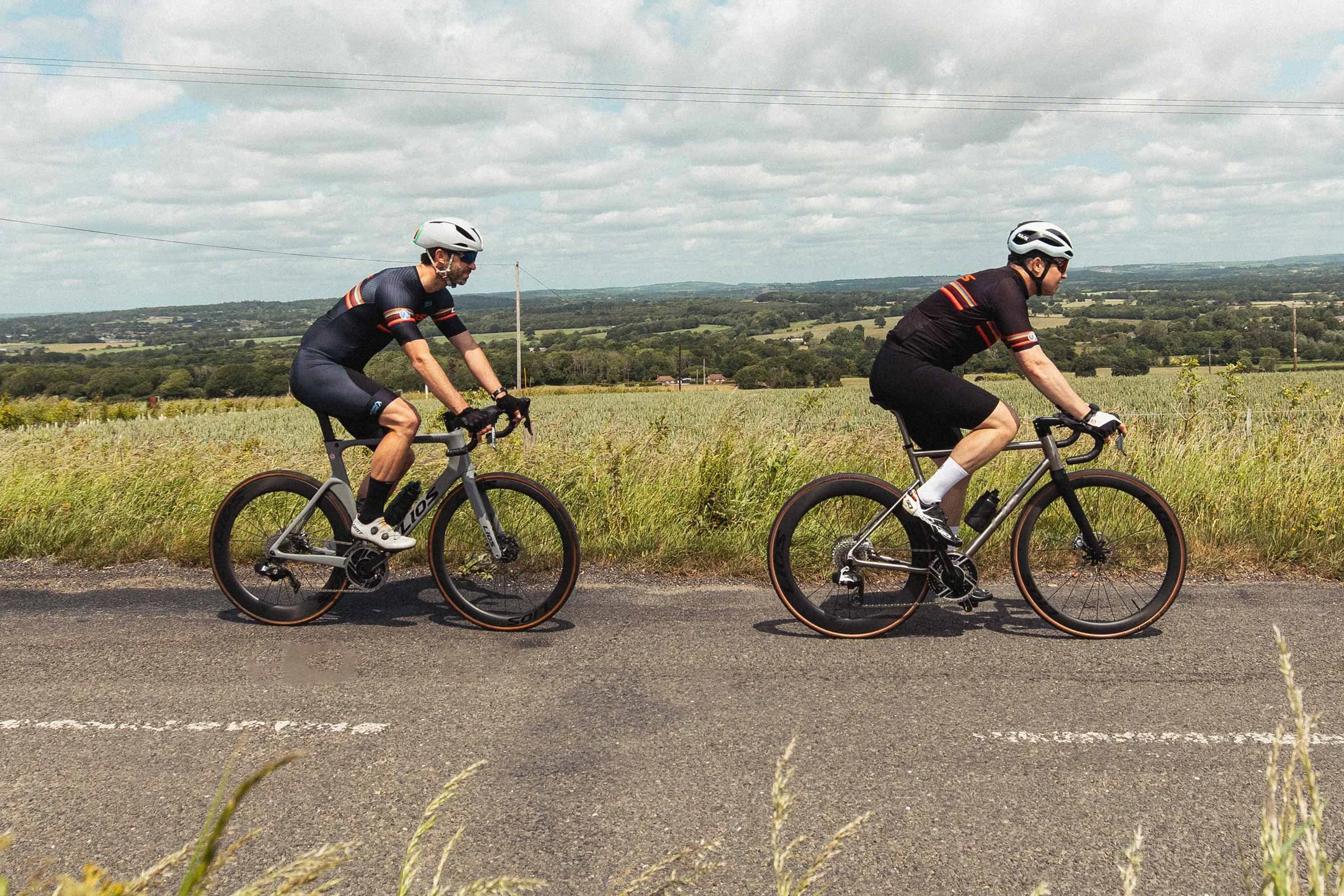 Photograph of two cyclists riding left to right along a country lane. The cyclist in front is riding a LIOS Titanium Road Race bike and the second cyclist is riding a LIOS Exactor carbon aero road bike.