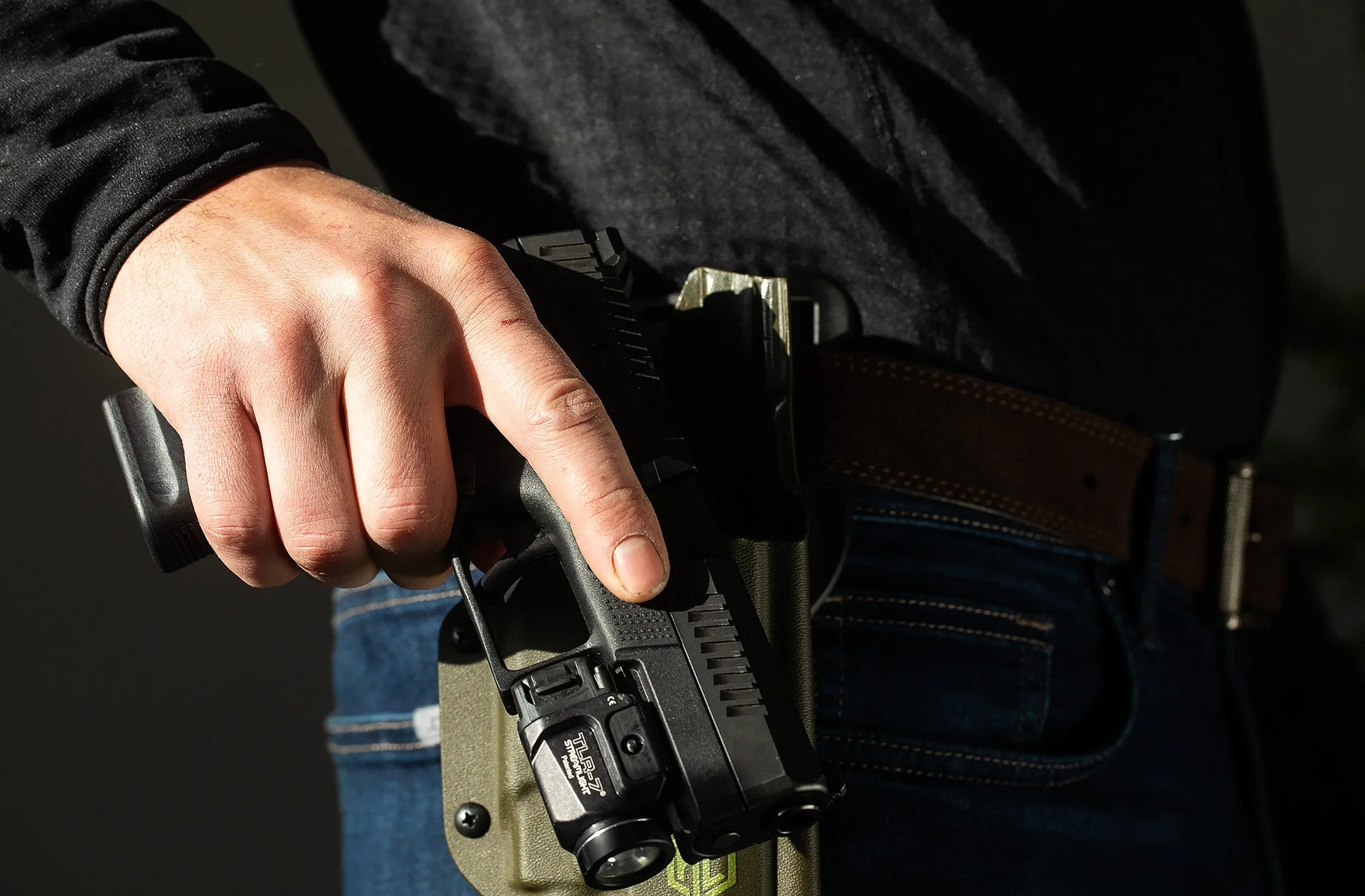 Fingers clasp the grip of a nine-millimeter handgun, displayed outside a hard plastic holster on a belt.