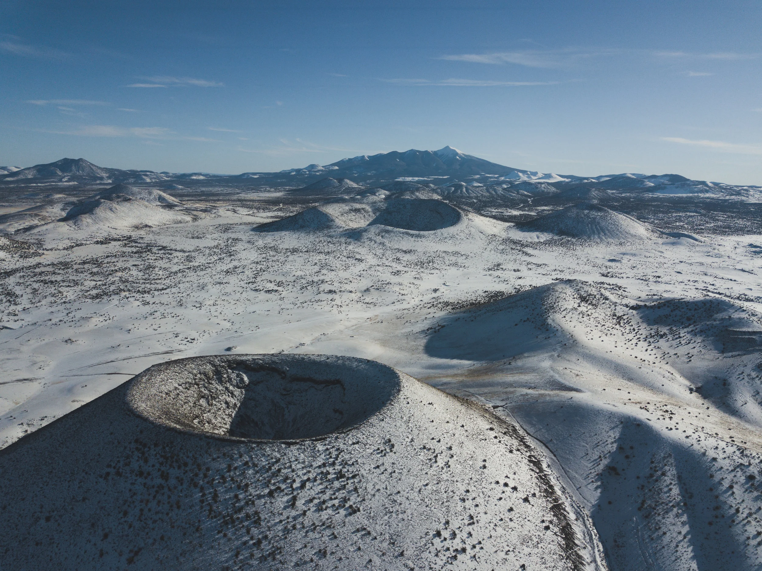 SP Crater in the foreground is a cinder cone which erupted between 60k - 71k years ago, its name is an abbreviation for Shit Pot. In the 1880s Rancher C.J. Babbit described its appearance as that of a chamber pot which over-flowed, not pictured here is the 4-mile long lava extending out from its base toward the viewer. Colton Crater is in the middle ground, it was one of the sites used for vehicle testing in the Apollo missions. These cinder cones, and 600 other cones, lava domes, and lava flows make up the San Francisco Volcanic field, named after the peaks that dominate the background of this image.