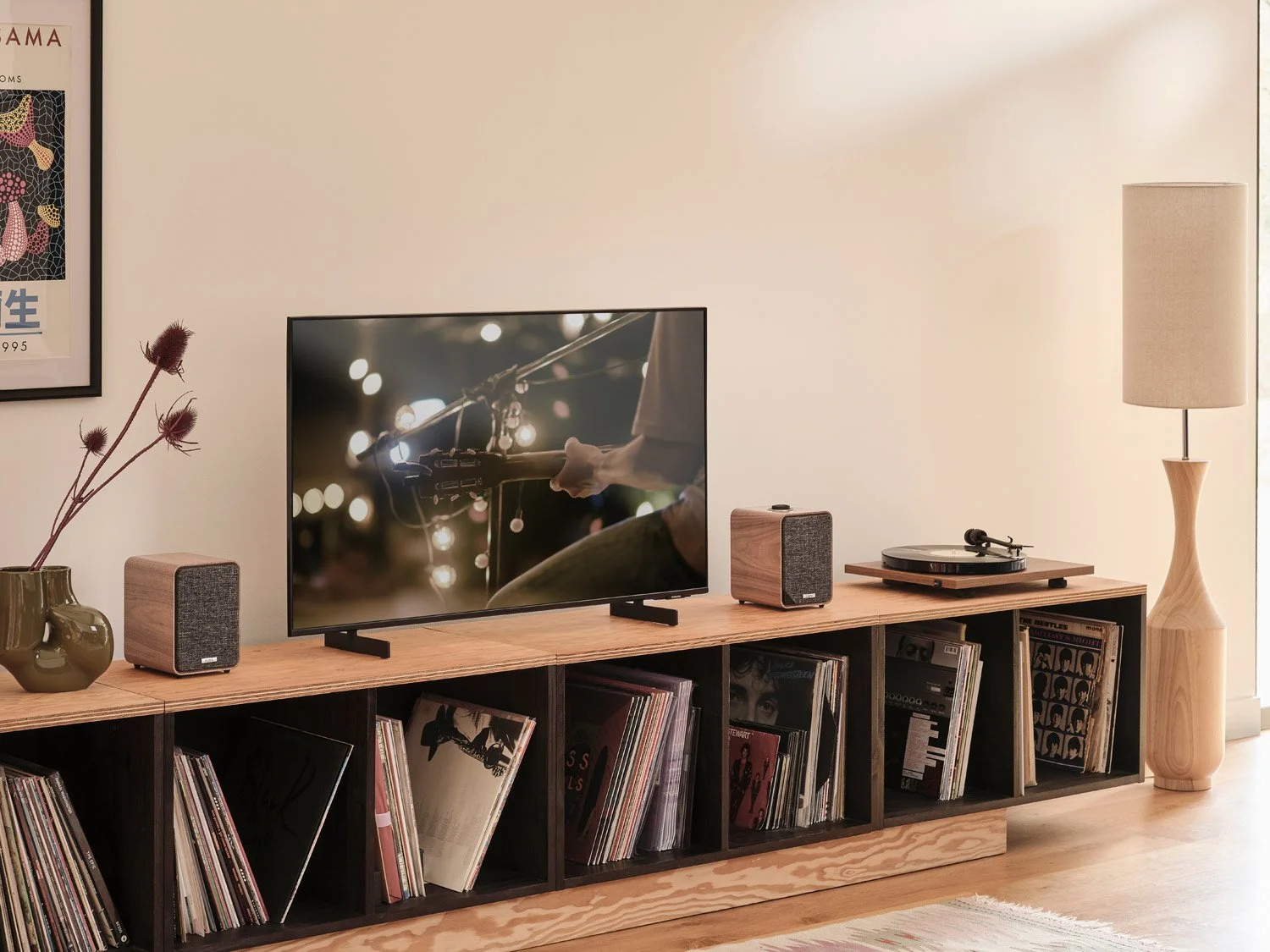 Living room entertainment setup featuring a flat-screen TV on a wooden media console, with two small speakers, a record player, vinyl records, a vase with dried flowers, and a tall wooden floor lamp next to a window.