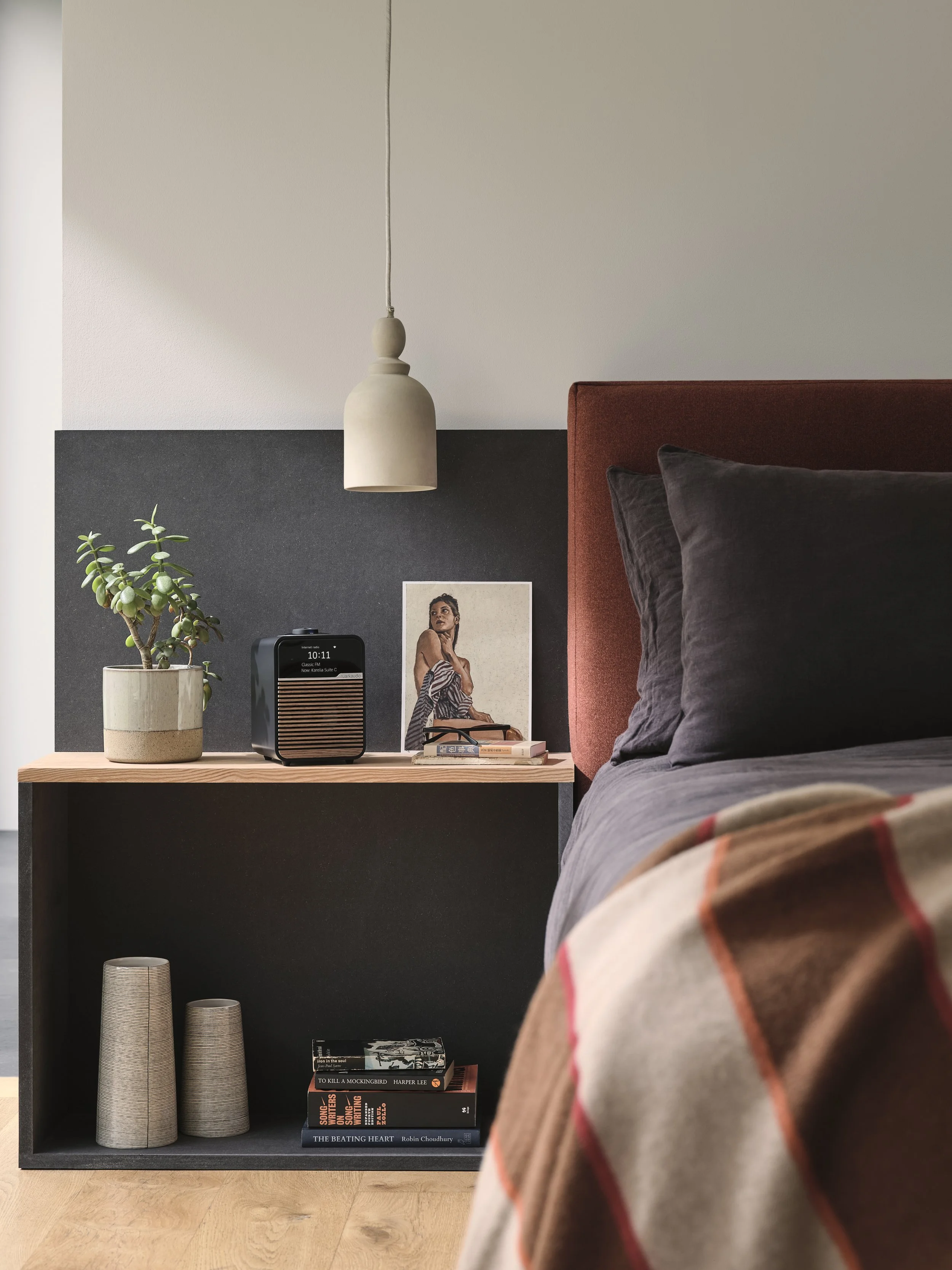 Close-up of a bedroom nightstand with a potted succulent, a black digital clock, a framed picture of a woman, a small speaker, and some books. The bed has dark gray sheets, black pillows, and a red headboard. A white hanging lamp is above the nightst
