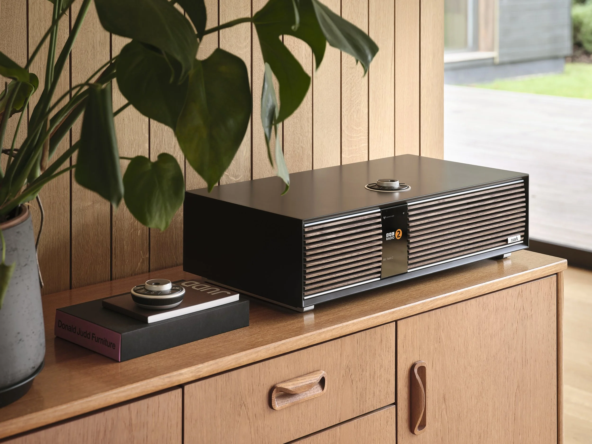 A vintage radio with a black and wood finish on a wooden sideboard, next to a potted plant, and a small black box with a circular object on top.