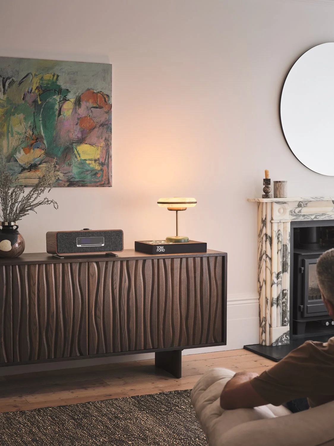 Living room with a wooden sideboard, a modern lamp, and a vintage speaker on top, a colorful abstract painting on the wall, a marble fireplace with candles and decor, a round mirror, and a person sitting on a couch.