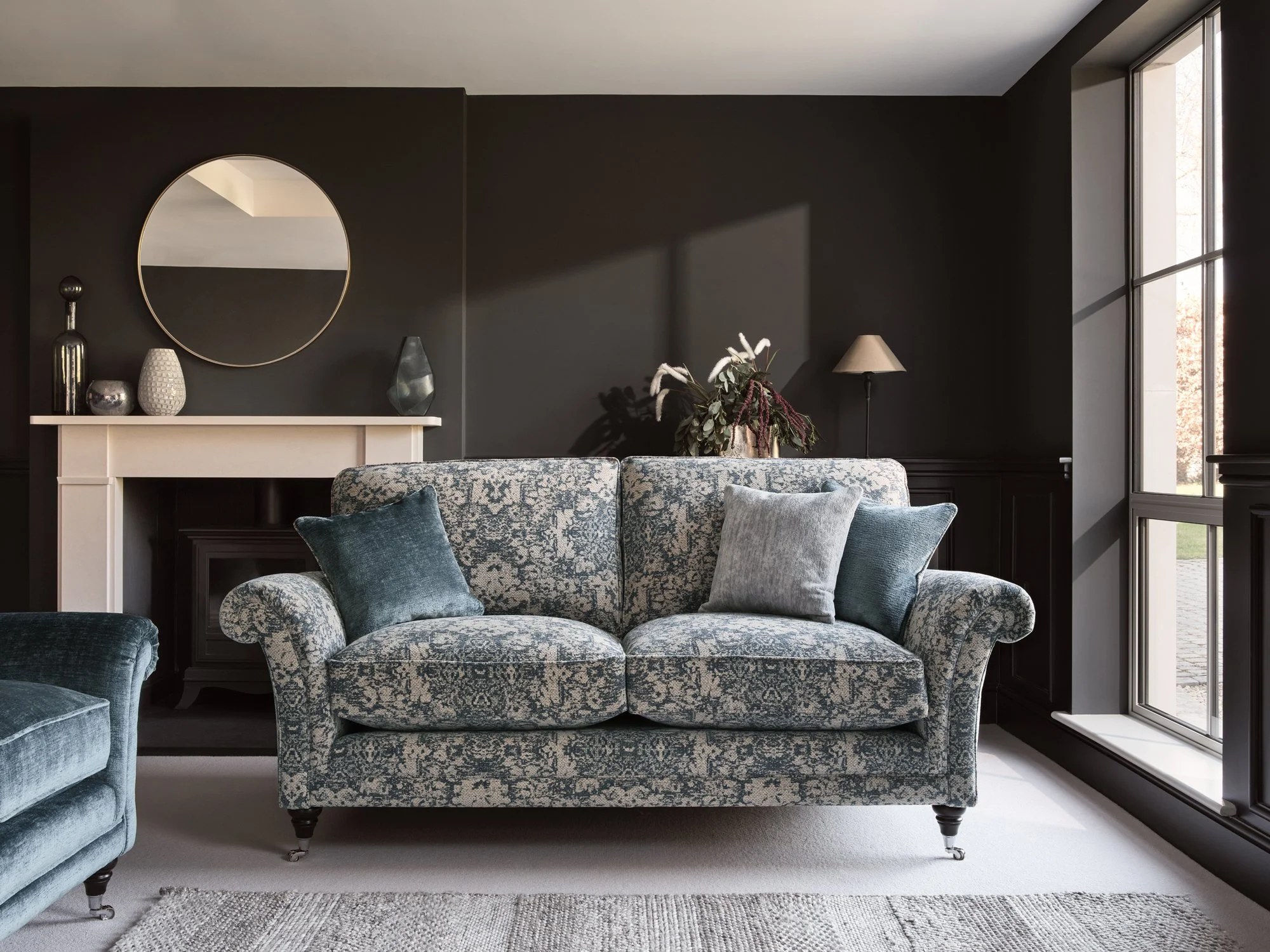 Living room with dark gray wall, patterned loveseat with blue and gray pillows, a side chair, a mantel with decorative vases, a round mirror, a flower arrangement, a floor lamp, and large windows.