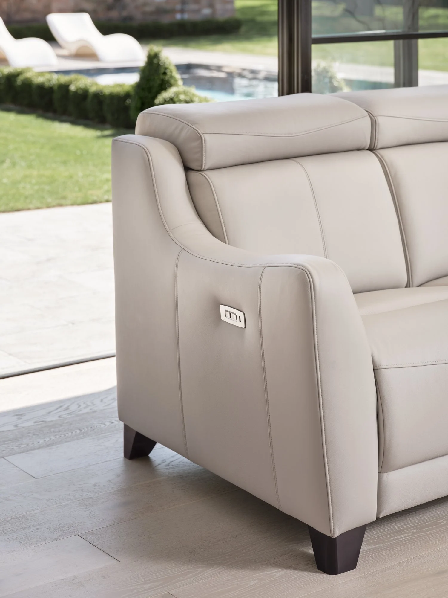 Close-up of a beige leather sofa with a power recline button on the side, situated inside near large glass windows that reveal a backyard with a pool and outdoor furniture.