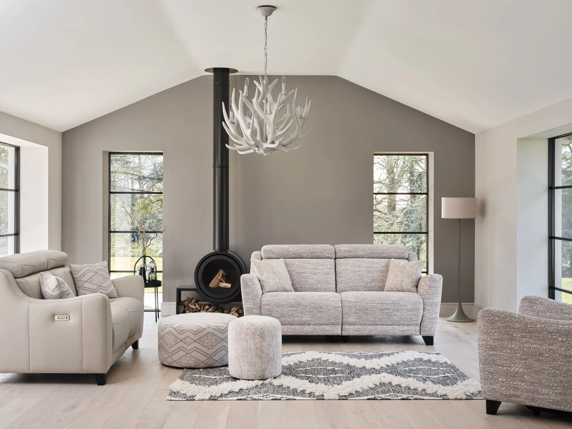 Modern living room with a gray wall, large windows, a beige recliner, a gray sofa, a white ottoman, a black wood stove, a floor lamp, and a patterned rug.