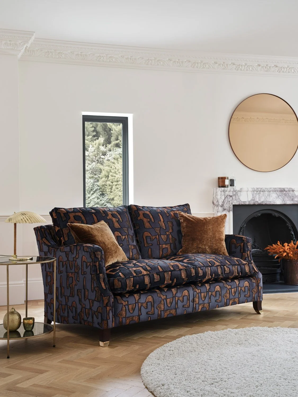 A living room with a patterned dark blue sofa, brown velvet pillows, a round side table with decorative items, a white shag rug, a fireplace with a marble mantel, a large circular mirror, and a window showing green trees outside.