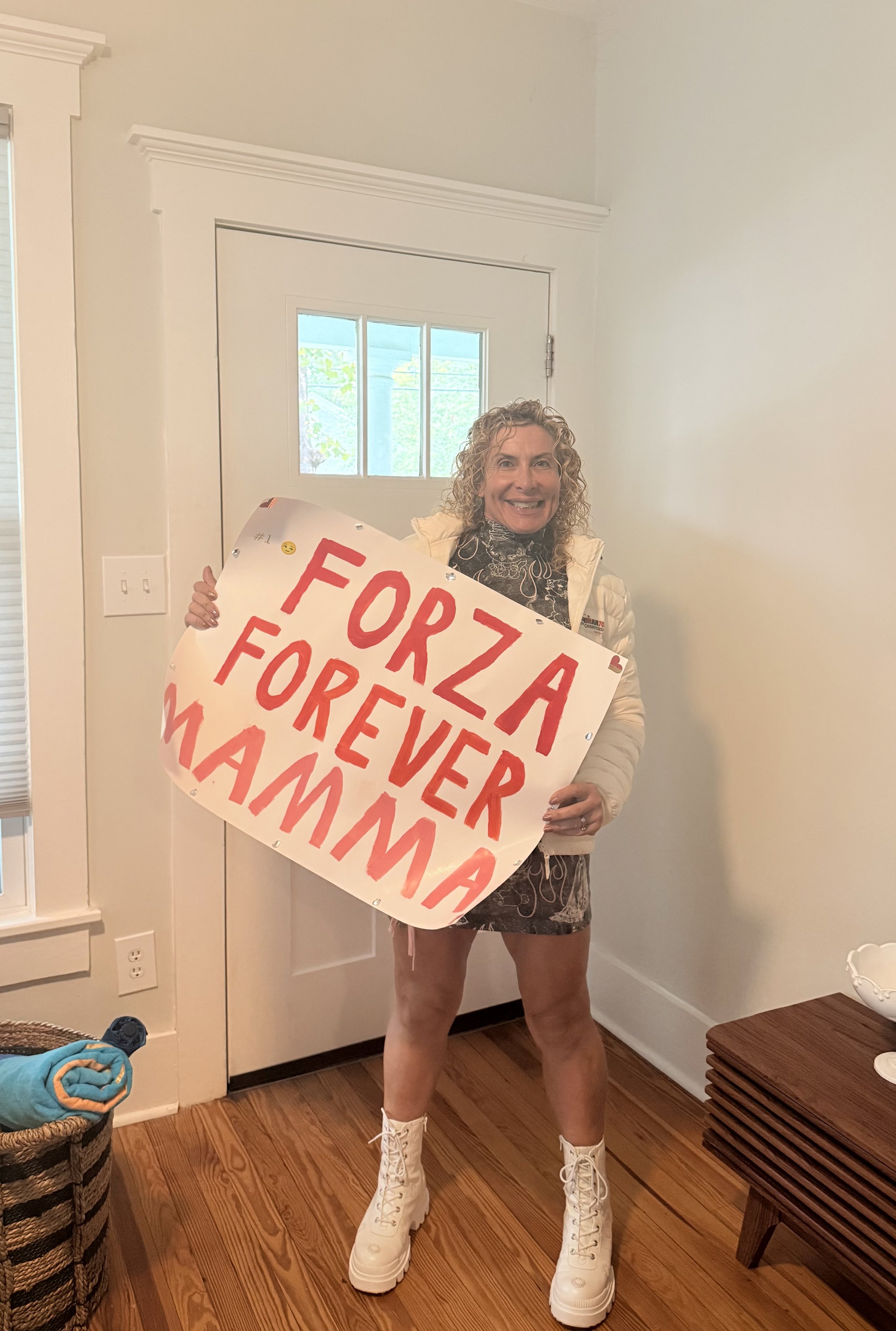 ForzaFit coach Rosa Laura smiling and holding the homemade “FORZA FOREVER MAMMA” sign made by her daughters Asia and Alessia during IRONMAN 70.3 North Carolina 2026.