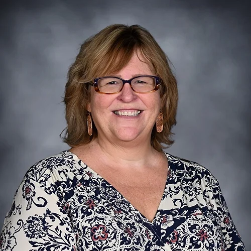 Person wearing glasses and a floral-patterned blouse, smiling against a gray background.