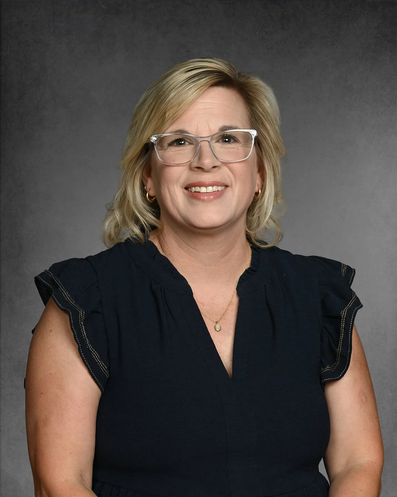 Woman with short blond hair wearing a black top and necklace, smiling against a gray background.
