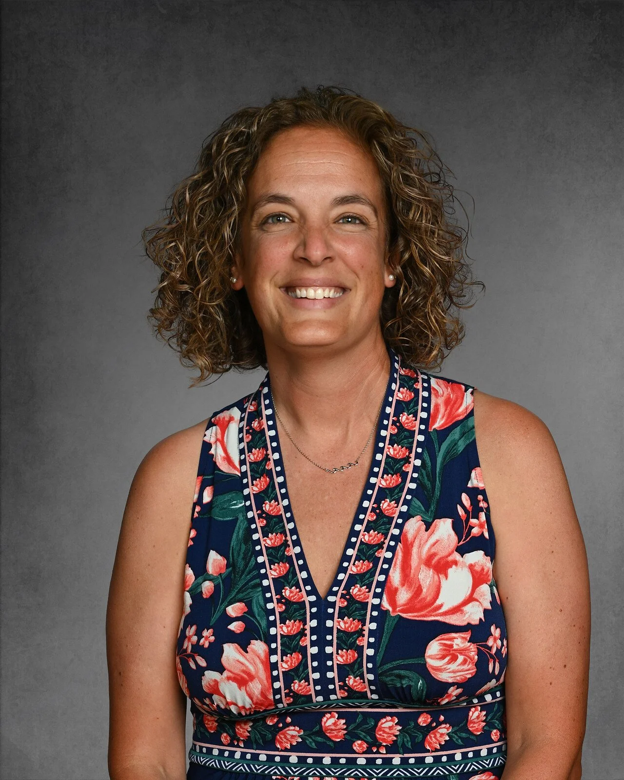 A woman with curly hair smiling, wearing a white sleeveless top with gray and black stripes, standing against a gray background.