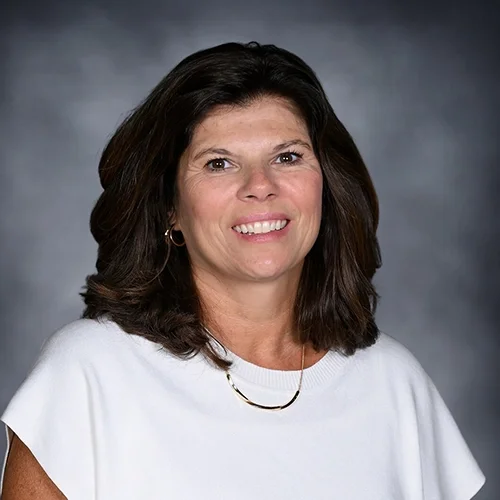 A woman with shoulder-length brown hair smiling, wearing a black sleeveless top and a necklace, posed against a dark gray background.