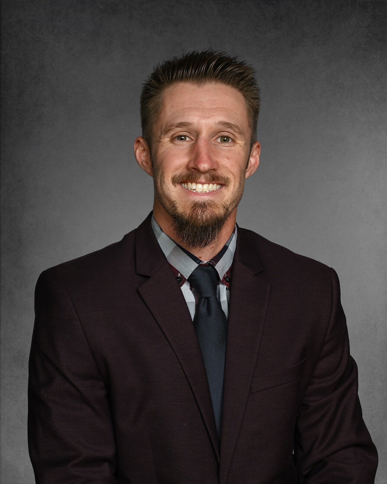 Man with a beard wearing a black suit, white shirt, and black tie, smiling at the camera.