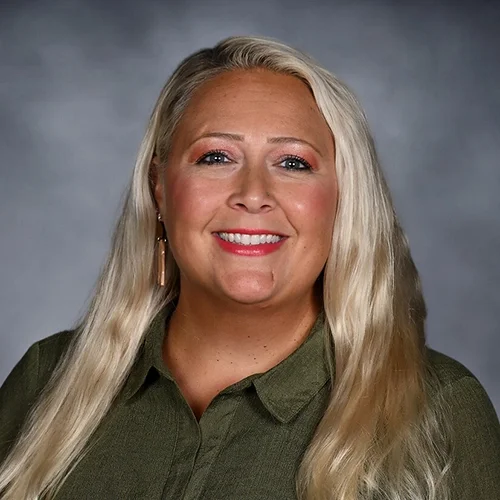 A woman with long, wavy blonde hair smiling, wearing a colorful floral blouse against a dark gray background.