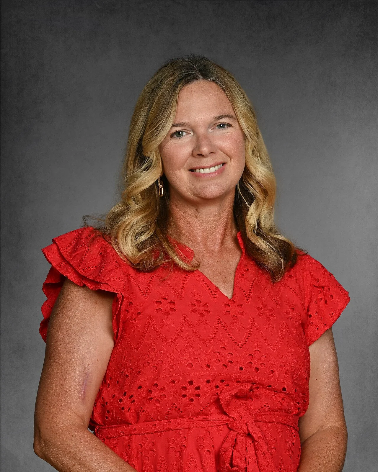 Smiling woman with blonde hair wearing a blue and white patterned blouse, posed against a gray background.