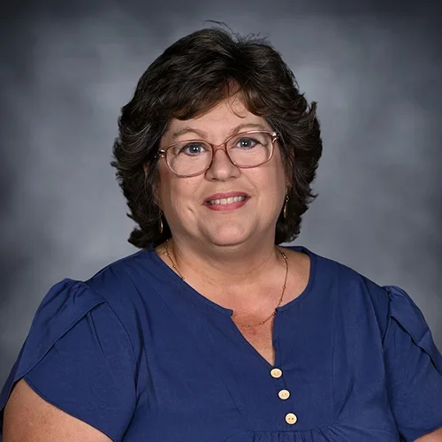 Woman with glasses smiling, wearing a striped blouse, against a dark background.