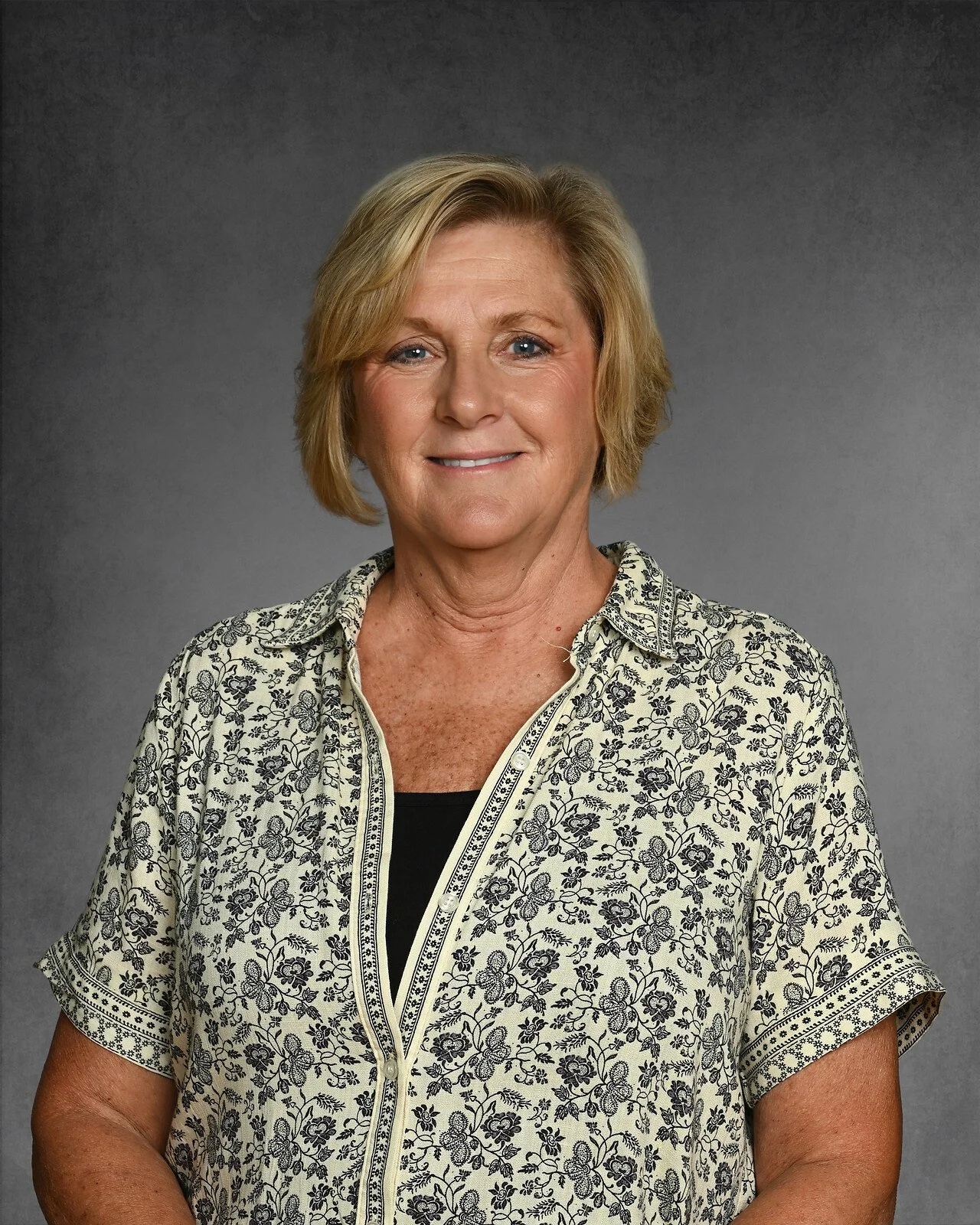 A smiling middle-aged woman with blonde hair in a navy blue dress, posing against a gray background.