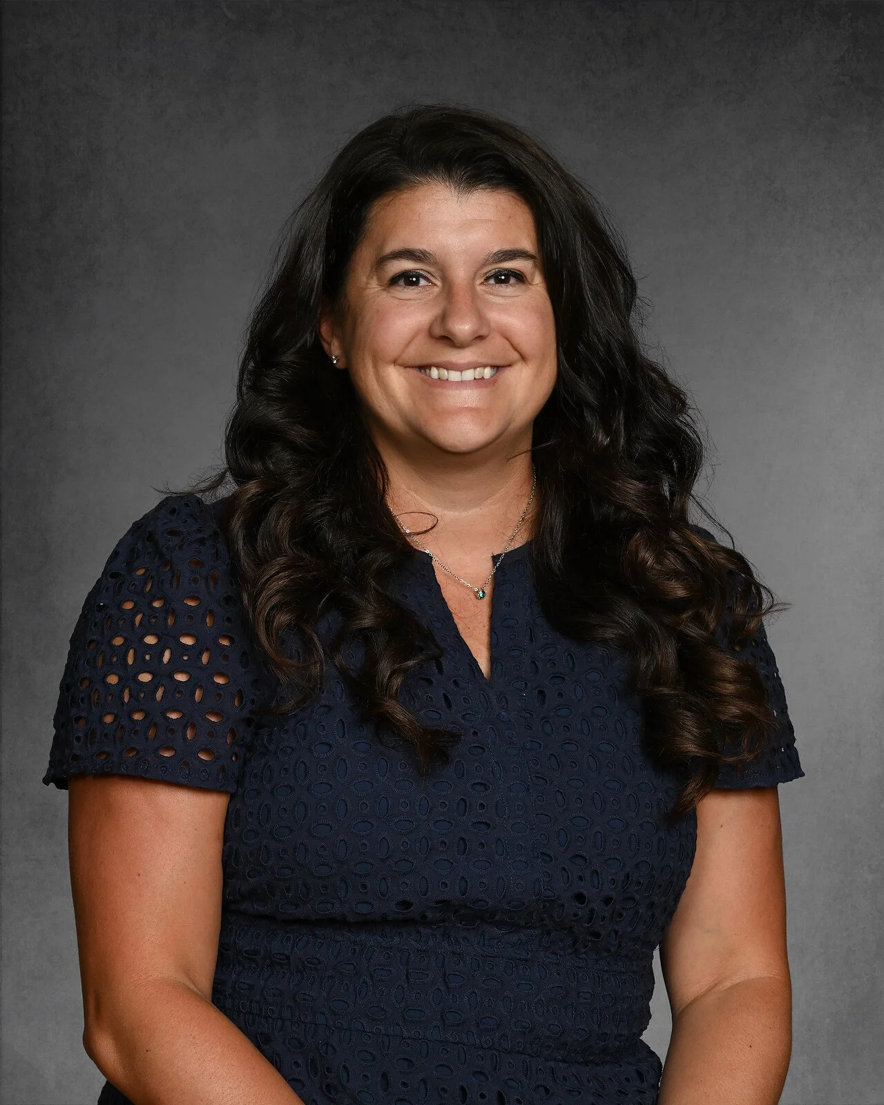 Woman with long brown hair wearing a floral-patterned top, smiling in front of a gray background.