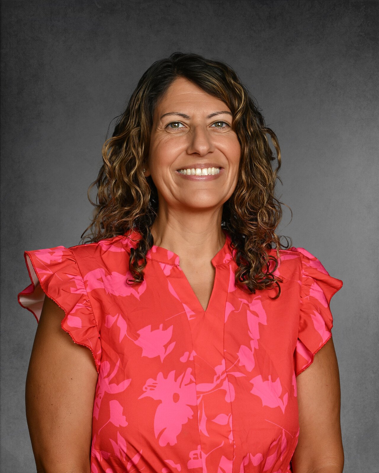 Smiling woman with curly hair wearing a floral blouse against a gray background.