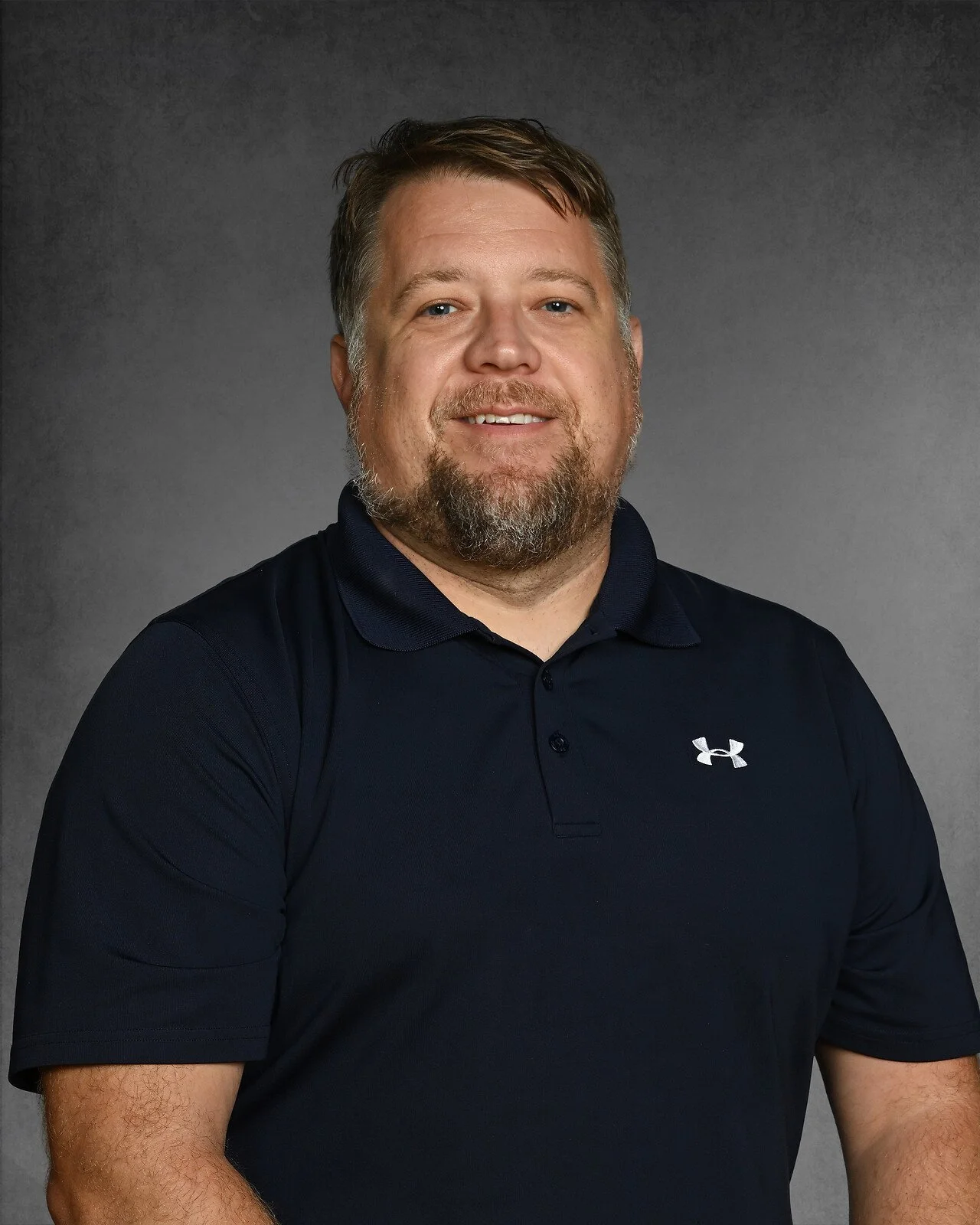 A man with light skin, red hair, and a beard, wearing a light blue button-up shirt and a white t-shirt, smiling at the camera against a dark gray background.
