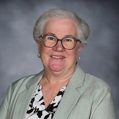 Headshot of an older woman with short, gray hair wearing glasses, a black blazer, and a patterned blouse, smiling against a dark gray background.