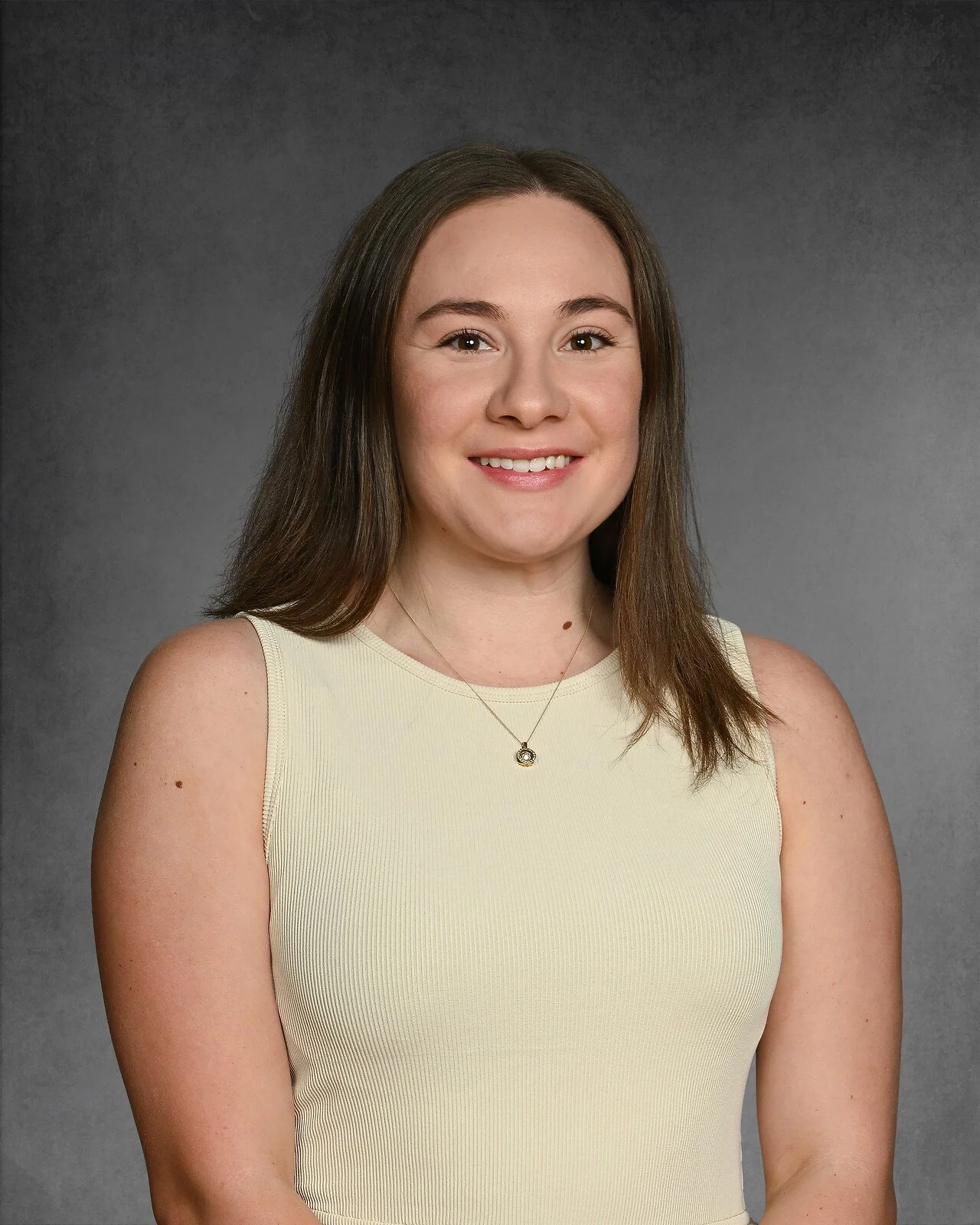 A young woman with long brown hair, smiling, wearing a floral sleeveless top and a necklace against a dark background.