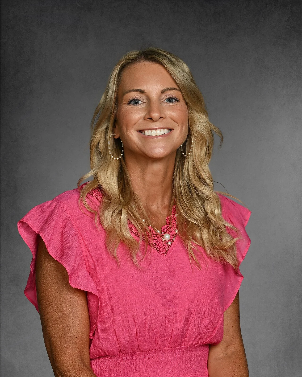 Smiling woman with long blonde hair, wearing a black top and necklace, against a gray background.