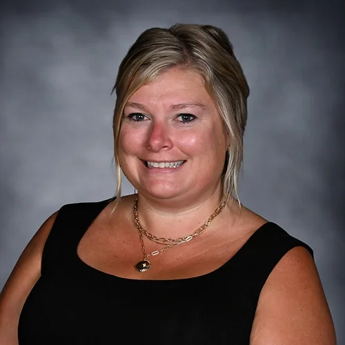 A woman with blonde hair, smiling, wearing large pearl earrings and a sleeveless black top, posing against a gray background.
