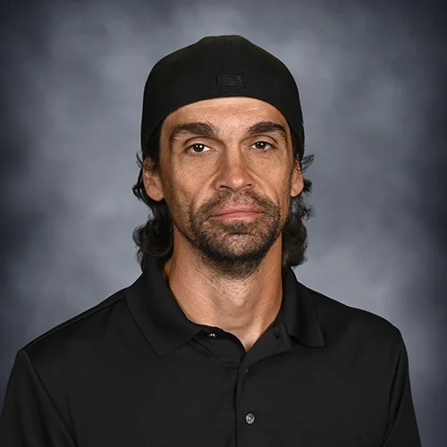 A man with long dark hair, a beard, wearing a black shirt and black cap, standing against a dark gray background.