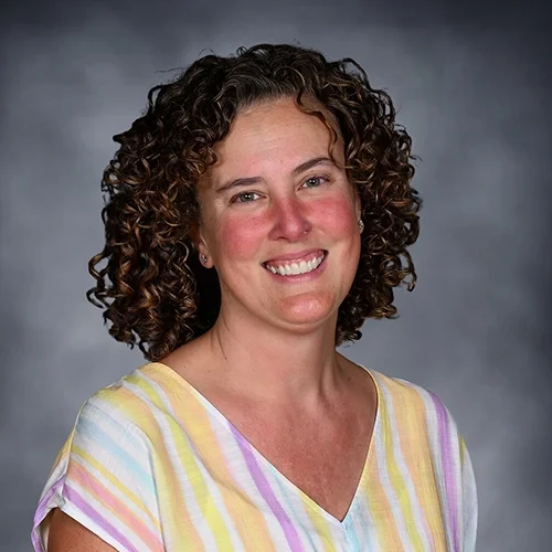 A woman with curly hair wearing a white sleeveless top and smiling against a gray background.