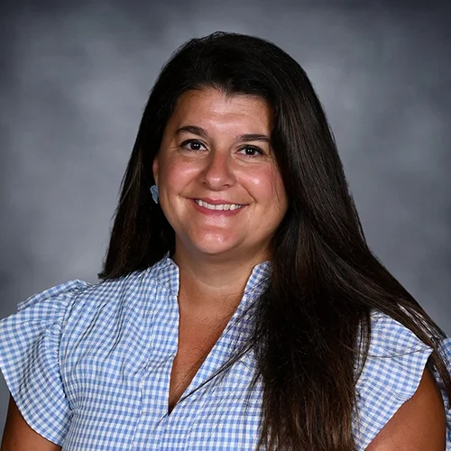 Woman with long brown hair wearing a floral-patterned top, smiling in front of a gray background.