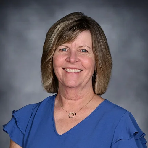 A woman with shoulder-length light brown hair smiling, wearing a blue blouse, against a dark gray background.