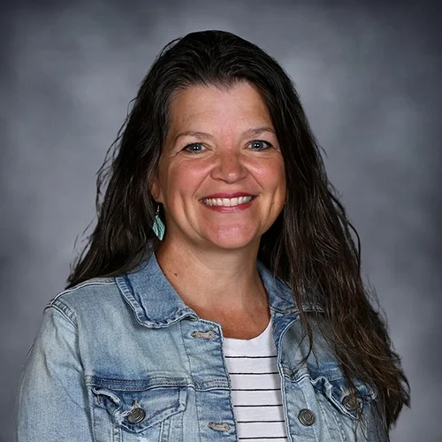 A woman with long, dark wavy hair smiling at the camera, wearing a cream-colored top with leopard print sleeves and a black beaded necklace with a large pendant.