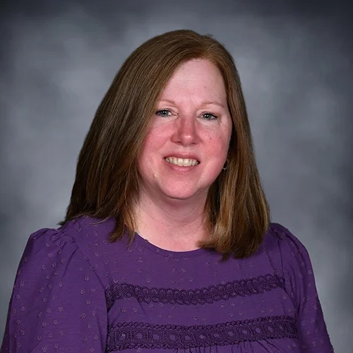 A woman with shoulder-length hair wearing a polka dot blouse, smiling against a gray background.