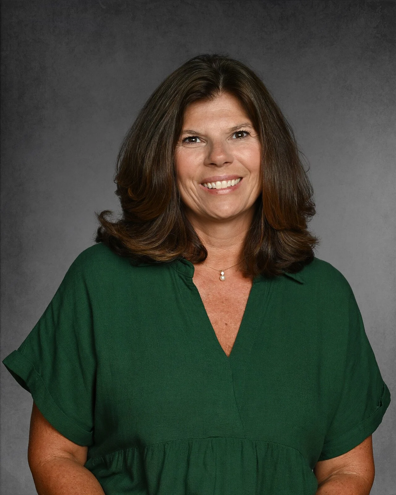 A woman with shoulder-length brown hair smiling, wearing a black sleeveless top and a necklace, posed against a dark gray background.
