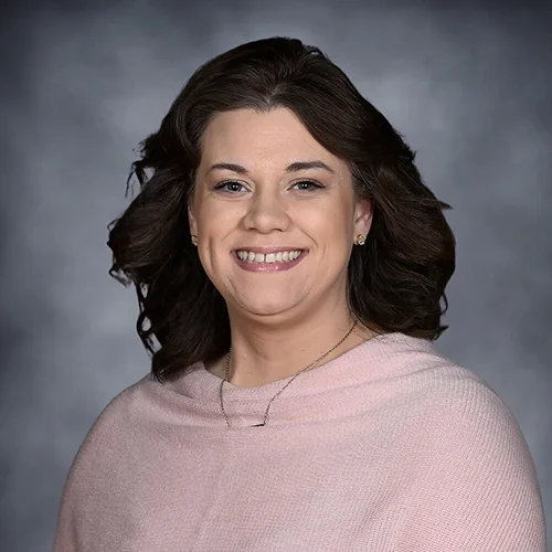 A woman with shoulder-length wavy blonde hair and bangs, smiling, wearing a black top and a necklace with a small pendant, against a gray background.