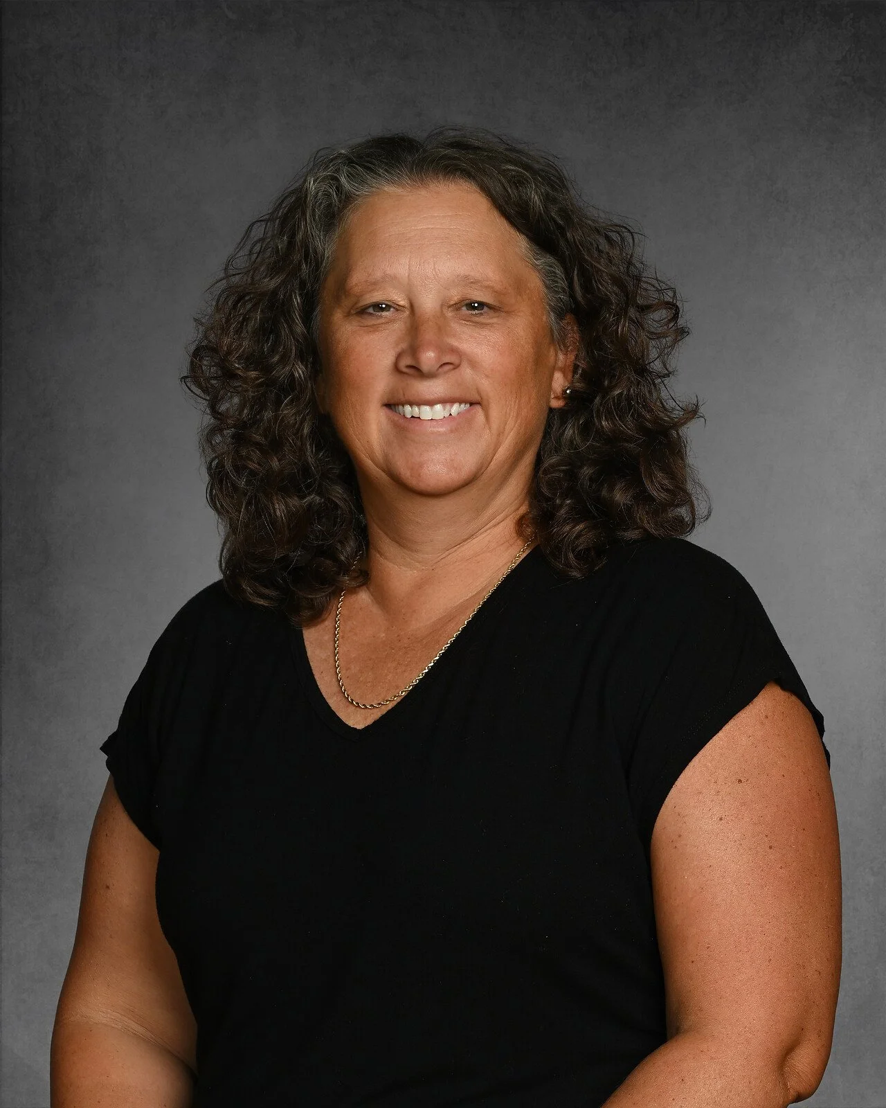 A woman with long curly brown hair, wearing a black polo shirt with an emblem that reads Holly Grove Christian School Athletics, standing against a dark gray background.