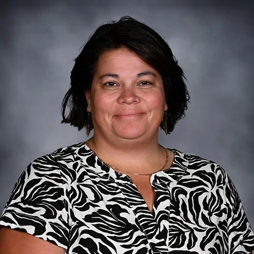A woman with short dark hair smiling, wearing a black and white patterned top, standing against a gray background.