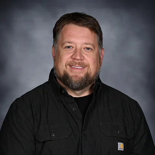 A man with light skin, red hair, and a beard, wearing a light blue button-up shirt and a white t-shirt, smiling at the camera against a dark gray background.