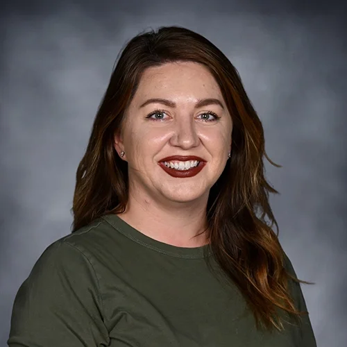 Portrait of a woman with shoulder-length brown hair, wearing a white top, smiling, against a gray background.
