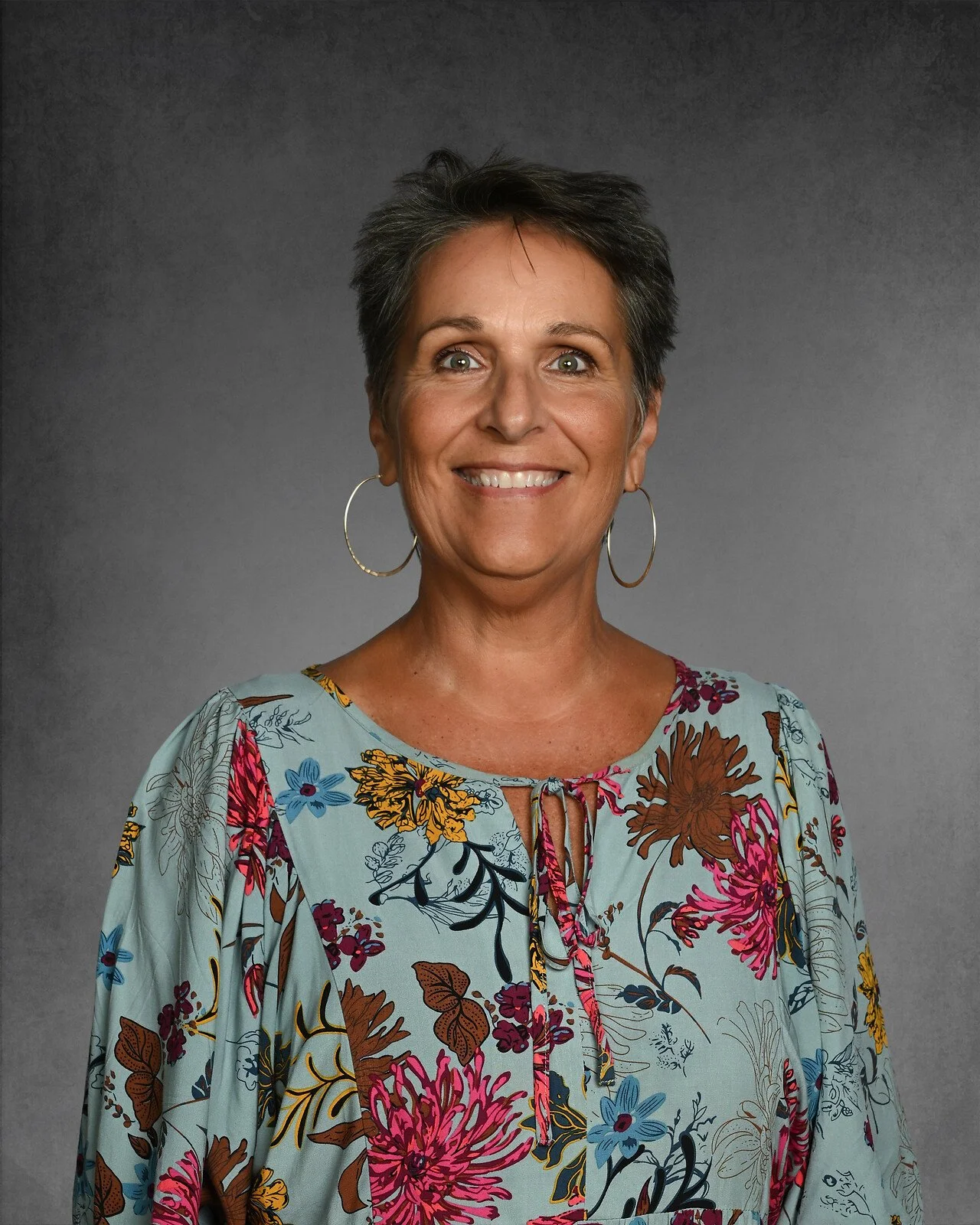 Smiling woman with short dark hair, wearing a white blouse and a patterned vest, with earrings, against a gray background.