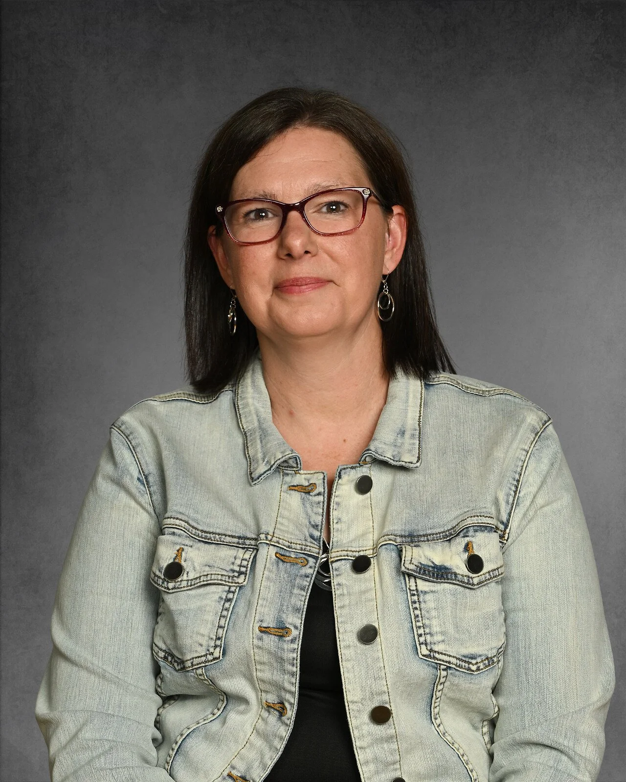 A woman with curly brown hair, glasses, and wearing a black sleeveless top, posed against a dark gray background.