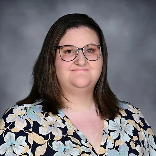 A woman with shoulder-length brown hair, smiling, wearing a red dress with white polka dots, against a gray background.