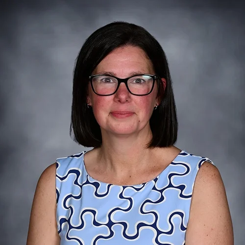 A woman with curly brown hair, glasses, and wearing a black sleeveless top, posed against a dark gray background.